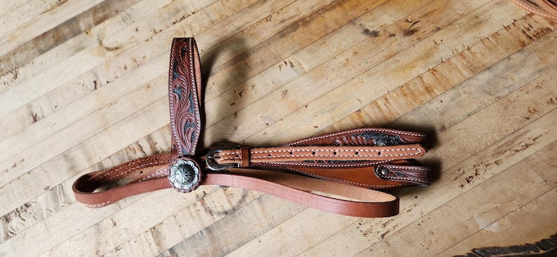 May include: A brown leather horse bridle with intricate floral tooling and a silver-colored concho. The bridle features multiple straps, a buckle, and a browband. The background is a wooden surface.