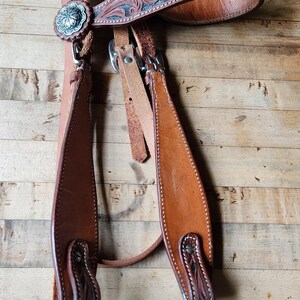 May include: A brown leather horse bridle with decorative tooling and silver-toned accents. The bridle features a floral design on the browband and cheek pieces. The leather is stitched with white thread.