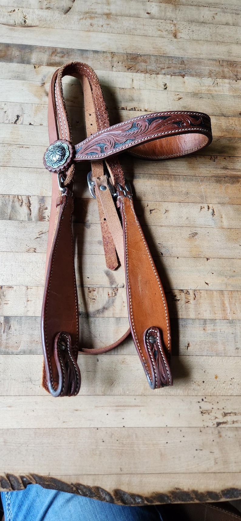 May include: A brown leather horse bridle with decorative tooling and silver-toned accents. The bridle features a floral design on the browband and cheek pieces. The leather is stitched with white thread.