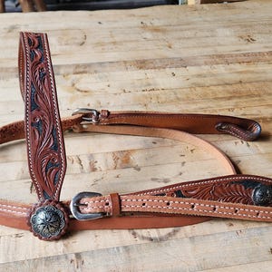 May include: A brown leather horse bridle with intricate floral tooling and silver-tone conchos. The bridle features a browband, cheek pieces, and a throatlatch, all connected with buckles. The leather is a medium brown color, with darker accents in the tooled areas.