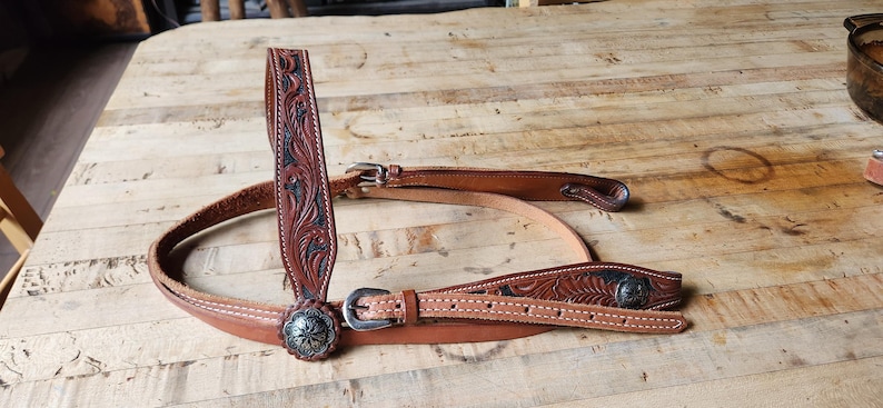 May include: A brown leather horse bridle with intricate floral tooling and silver-tone conchos. The bridle features a browband, cheek pieces, and a throatlatch, all connected with buckles. The leather is a medium brown color, with darker accents in the tooled areas.