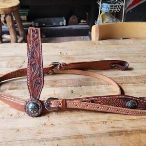 May include: A brown leather horse bridle with intricate floral tooling and silver-toned conchos. The bridle features a browband, cheek pieces, and a throat latch, all connected with buckles. The leather is a rich, warm brown color.