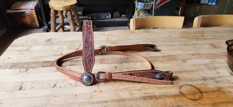 May include: A brown leather horse bridle with intricate floral tooling and silver-toned conchos. The bridle features a browband, cheek pieces, and a throat latch, all connected with buckles. The leather is a rich, warm brown color.