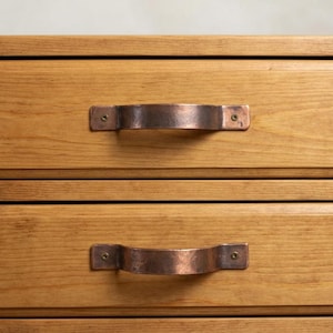 May include: A light brown wooden chest of drawers featuring two stacked drawers. Each drawer is equipped with a horizontal, hammered copper-toned metal handle secured by two small screws on either side of the handle base.