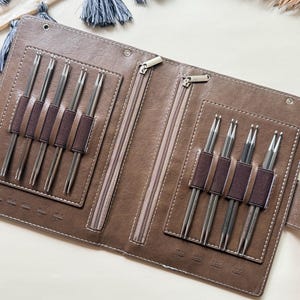 May include: A brown faux leather folding case containing eight silver-toned metal knitting needles held by dark brown elastic bands. The case features two zippered compartments and embossed size markings in inches and millimeters for each needle.