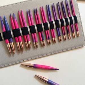 Puede incluir: Un juego de agujas de tejer intercambiables con ejes de madera veteada en rosa y morado y puntas de color cobre metálico. Las agujas están organizadas en pares dentro de un estuche de tela gris claro con elásticos negros, y dos agujas sueltas.