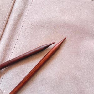 Könnte beinhalten: Zwei dunkelbraune Stricknadeln aus Holz mit spitz zulaufenden Enden liegen auf einem hellrosa Etui aus wildlederartigem Stoff. Eine Nadel hat einen goldfarbenen Metallsockel, die andere besteht aus massivem Holz. Das Etui hat sichtbare Nähte.