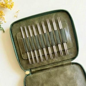 Könnte beinhalten: Ein olivgrünes Etui mit Reißverschluss, das acht doppelseitige Stricknadeln aus Metall enthält. Die Nadeln werden durch einzelne elastische Schlaufen im Inneren des Etuis gehalten, das eine strukturierte, lederartige Oberfläche aufweist.