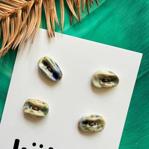 May include: Four oval ceramic buttons with a glossy finish in shades of cream, green, and blue, each featuring a central dark brown hole. The buttons are attached to a white display card with the black text "këkön" printed at the bottom.
