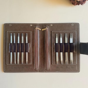 May include: A brown folding case containing nine silver-toned double-pointed knitting needles held in dark brown elastic loops. The case features two vertical zippered compartments in the center and white stitching along the edges of the interior.