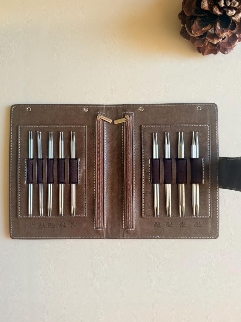 May include: A brown folding case containing nine silver-toned double-pointed knitting needles held in dark brown elastic loops. The case features two vertical zippered compartments in the center and white stitching along the edges of the interior.