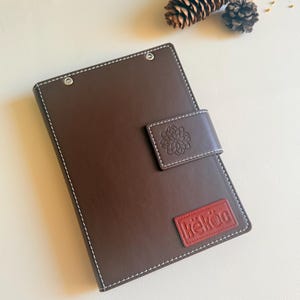 May include: A dark brown rectangular notebook with white contrast stitching, a magnetic closure flap featuring an embossed floral design, and a red rectangular patch at the bottom right corner with the white embossed text "k&euml;k&ouml;c".