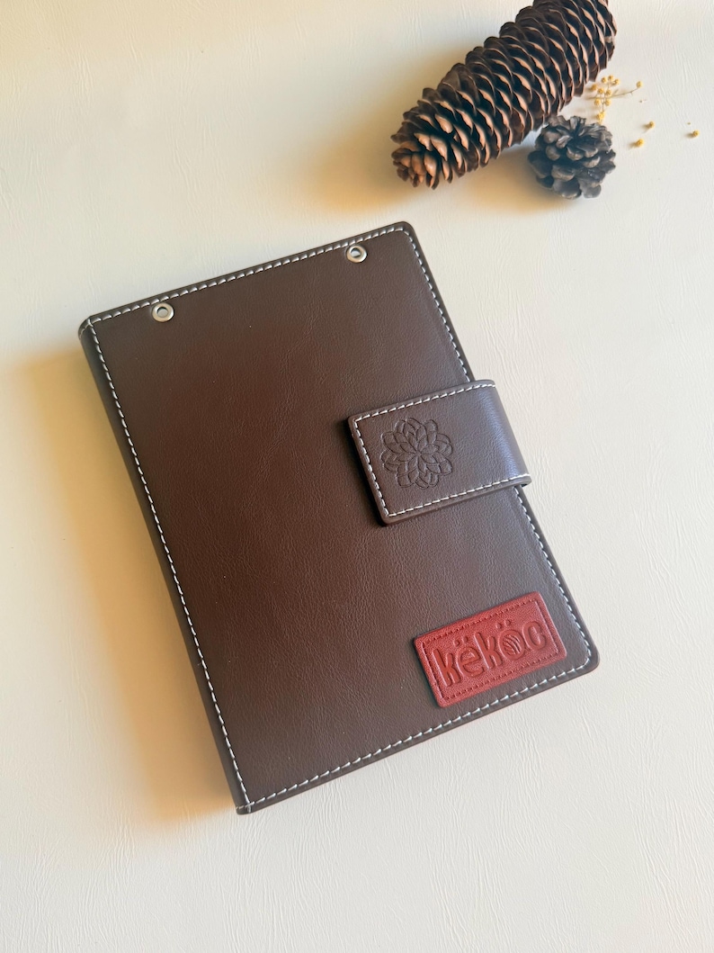 May include: A dark brown rectangular notebook with white contrast stitching, a magnetic closure flap featuring an embossed floral design, and a red rectangular patch at the bottom right corner with the white embossed text "k&euml;k&ouml;c".