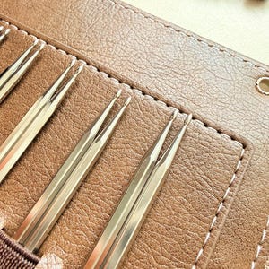 May include: A set of metallic knitting needles with rounded tips, organized in a tan textured case with white stitching. The needles are silver-toned and feature a smooth, reflective finish, held securely in individual slots within the brown case.