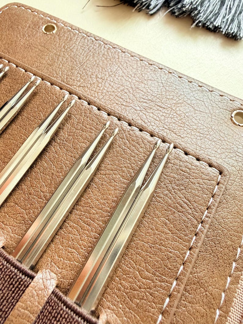 May include: A set of metallic knitting needles with rounded tips, organized in a tan textured case with white stitching. The needles are silver-toned and feature a smooth, reflective finish, held securely in individual slots within the brown case.
