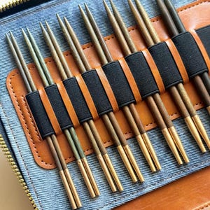 May include: A set of knitting needles with wooden shafts and metallic gold-toned tips, organized in a blue denim and tan leather case with black elastic bands. The needles vary in shade from light to dark brown and are arranged in pairs.