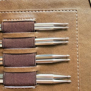 May include: A tan faux leather case with a zippered top compartment and a rectangular stitched panel featuring four dark brown elastic loops. Each loop holds two silver-toned metal double-pointed knitting needles with rounded tips.