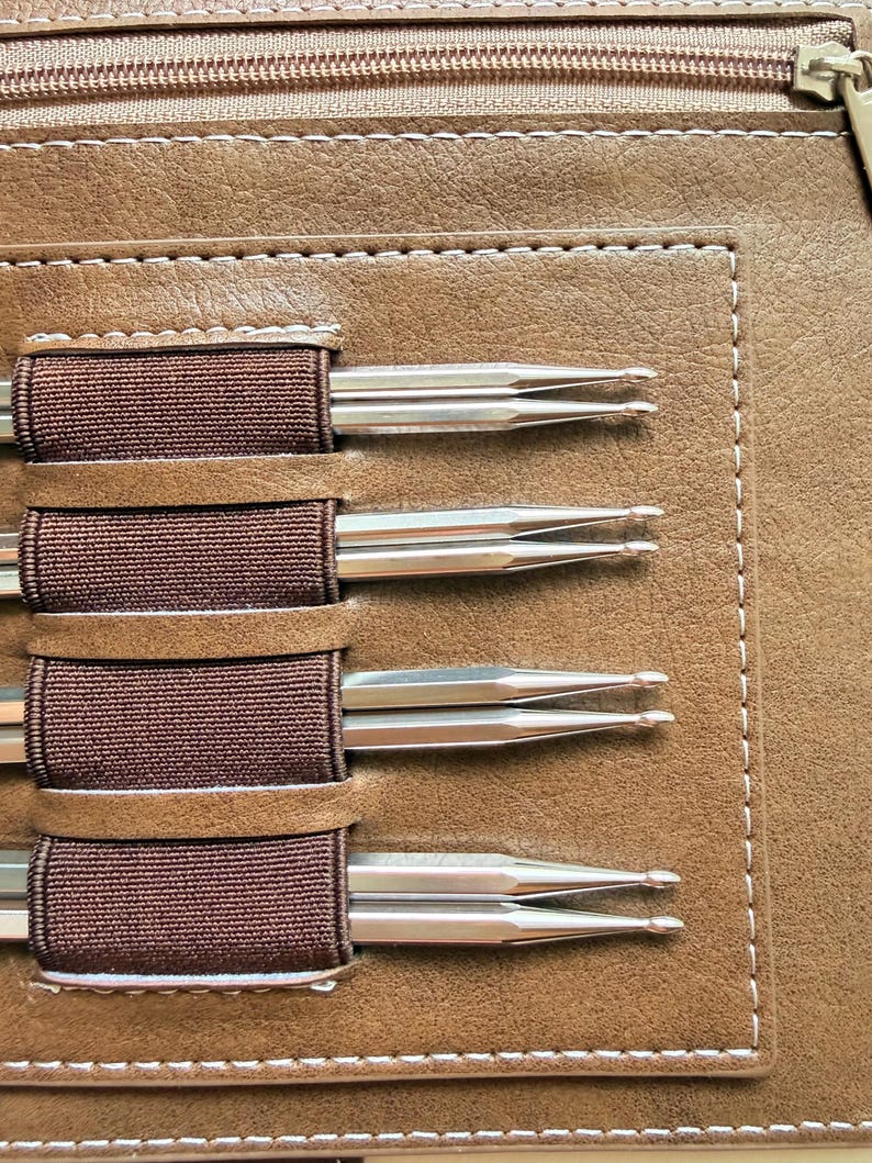 May include: A tan faux leather case with a zippered top compartment and a rectangular stitched panel featuring four dark brown elastic loops. Each loop holds two silver-toned metal double-pointed knitting needles with rounded tips.