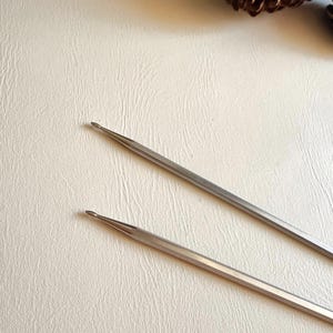 May include: Two silver-colored metal knitting needles with tapered, pointed tips. The needles are positioned diagonally across the frame, showing a smooth, reflective surface finish. A brown pinecone is visible in the upper right corner.