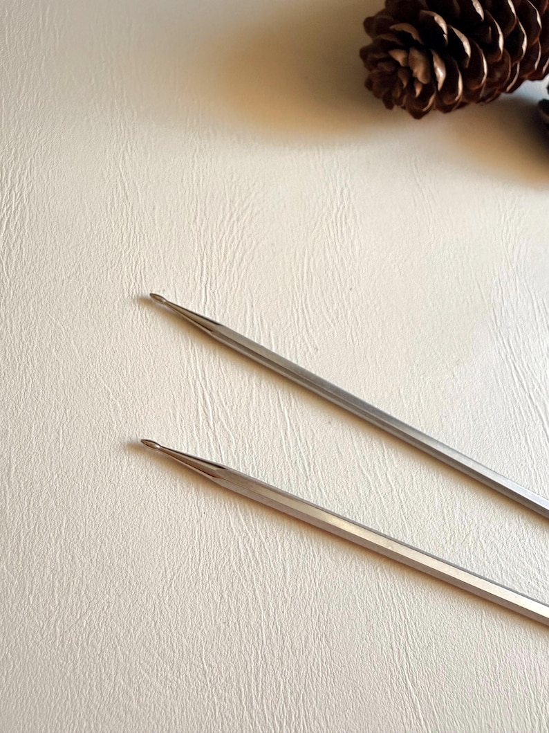 May include: Two silver-colored metal knitting needles with tapered, pointed tips. The needles are positioned diagonally across the frame, showing a smooth, reflective surface finish. A brown pinecone is visible in the upper right corner.