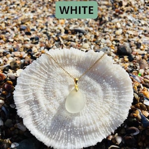 May include: A gold chain necklace with a teardrop-shaped, frosted white pendant. The necklace is displayed on a white coral-like object, set against a background of seashells and sand. The word "WHITE" is displayed in a green rectangle.