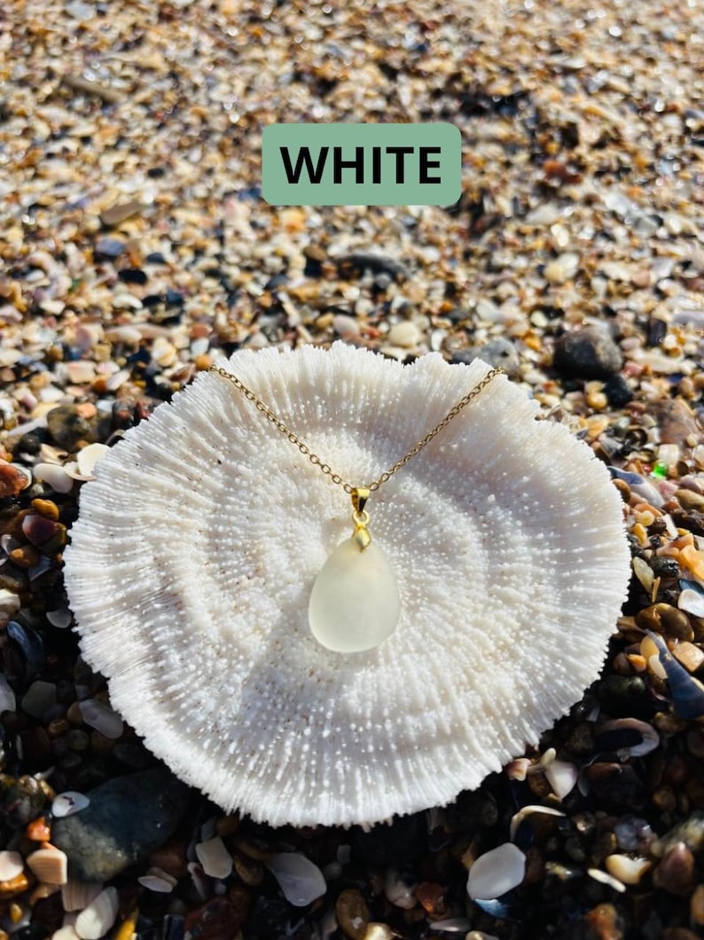May include: A gold chain necklace with a teardrop-shaped, frosted white pendant. The necklace is displayed on a white coral-like object, set against a background of seashells and sand. The word "WHITE" is displayed in a green rectangle.