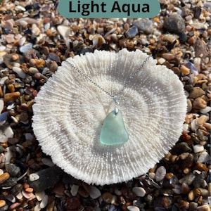 May include: A light aqua sea glass pendant necklace with a silver chain. The pendant is a triangular shape and rests on a white coral-like surface. The text "Light Aqua" is in a green rectangle.