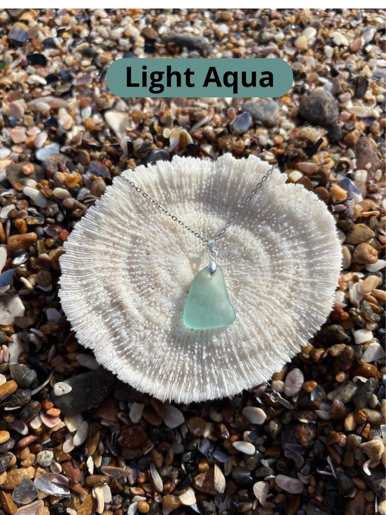 May include: A light aqua sea glass pendant necklace with a silver chain. The pendant is a triangular shape and rests on a white coral-like surface. The text "Light Aqua" is in a green rectangle.