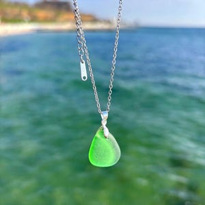 May include: A silver necklace with a bright green, teardrop-shaped pendant. The pendant is made of sea glass and hangs from a delicate chain. The background is a blurred view of the ocean and sky.