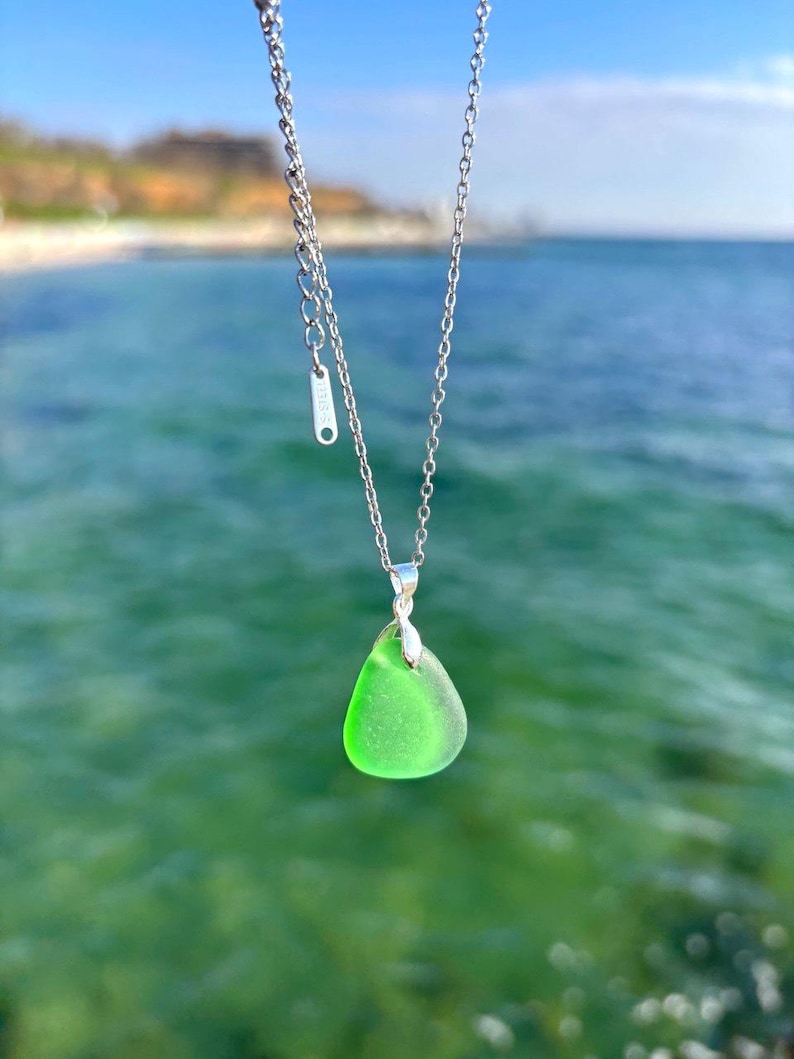 May include: A silver necklace with a bright green, teardrop-shaped pendant. The pendant is made of sea glass and hangs from a delicate chain. The background is a blurred view of the ocean and sky.