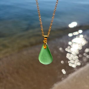 May include: A gold-colored chain necklace with a teardrop-shaped, seafoam green pendant. The pendant is smooth and translucent. The background shows a blurred view of the ocean and sand.