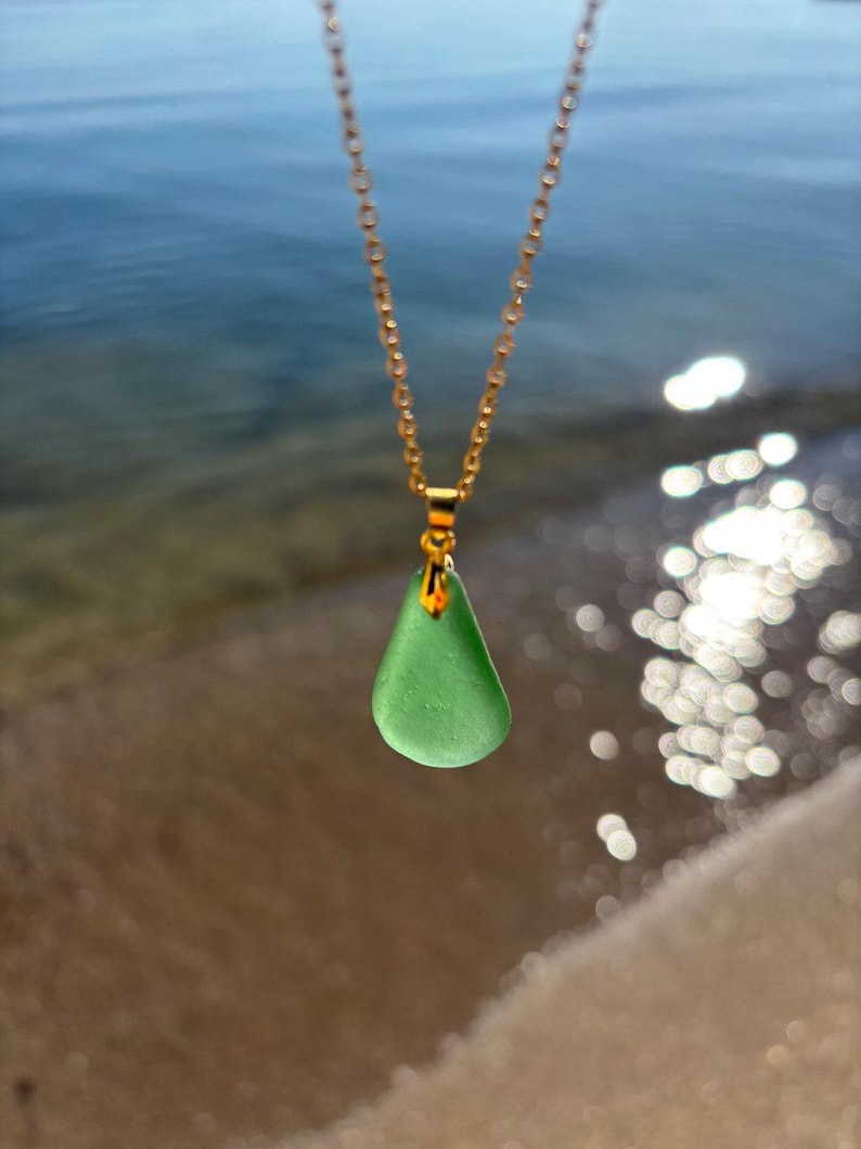 May include: A gold-colored chain necklace with a teardrop-shaped, seafoam green pendant. The pendant is smooth and translucent. The background shows a blurred view of the ocean and sand.