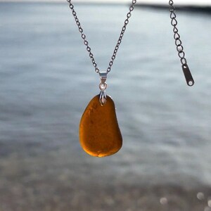 May include: A silver chain necklace with a pendant made of a smooth, amber-colored stone. The pendant is teardrop-shaped and hangs from a small silver bail. The background is a blurred view of the ocean.