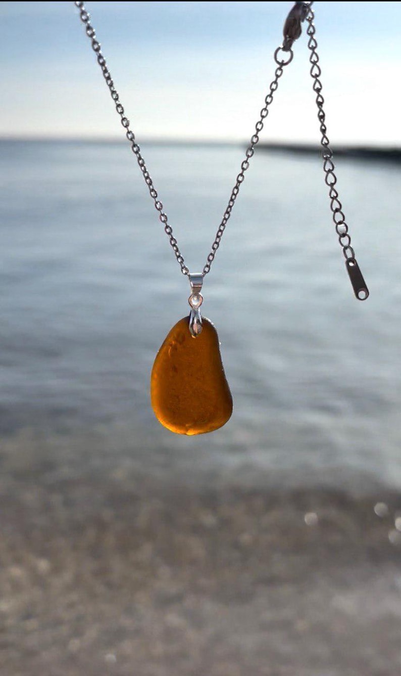 May include: A silver chain necklace with a pendant made of a smooth, amber-colored stone. The pendant is teardrop-shaped and hangs from a small silver bail. The background is a blurred view of the ocean.