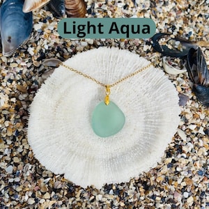 May include: A light aqua pendant necklace with a gold chain. The pendant is a smooth, irregular shape. The necklace is displayed on a white coral-like surface, with the text "Light Aqua" in a green rectangle.