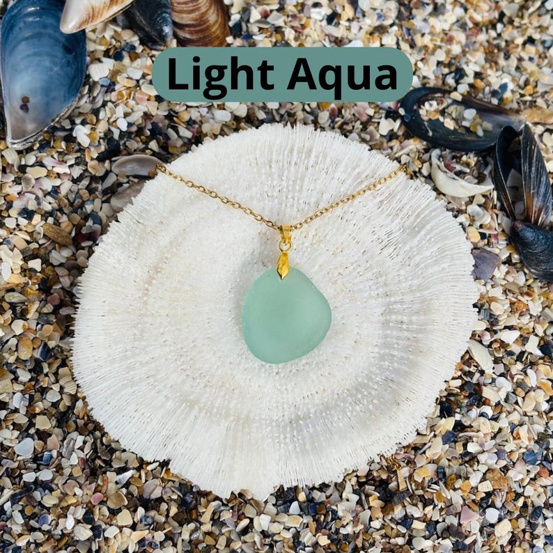 May include: A light aqua pendant necklace with a gold chain. The pendant is a smooth, irregular shape. The necklace is displayed on a white coral-like surface, with the text "Light Aqua" in a green rectangle.