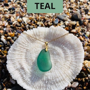 May include: A teal-colored sea glass pendant necklace with a gold chain. The pendant is a smooth, rounded shape. The necklace rests on a white coral-like surface, with the word "TEAL" in a green rectangle above it.