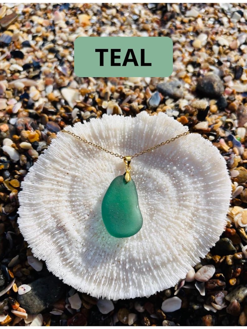 May include: A teal-colored sea glass pendant necklace with a gold chain. The pendant is a smooth, rounded shape. The necklace rests on a white coral-like surface, with the word "TEAL" in a green rectangle above it.