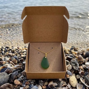 May include: A green teardrop pendant necklace with a gold chain is displayed inside a brown cardboard box. The box is open, revealing the necklace against a tan background. The box is on a beach with pebbles and shells.