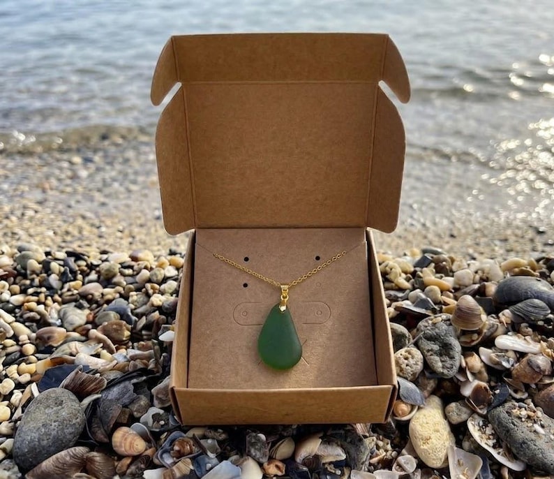 May include: A green teardrop pendant necklace with a gold chain is displayed inside a brown cardboard box. The box is open, revealing the necklace against a tan background. The box is on a beach with pebbles and shells.