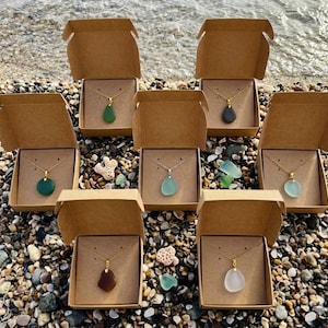 May include: Seven necklaces with gold chains and teardrop-shaped pendants in various colors, including green, blue, and brown, are displayed in open cardboard boxes. The boxes are arranged on a pebble beach near the water.