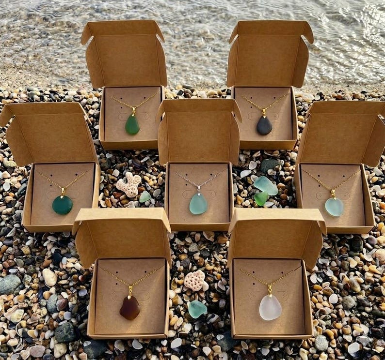 May include: Seven necklaces with gold chains and teardrop-shaped pendants in various colors, including green, blue, and brown, are displayed in open cardboard boxes. The boxes are arranged on a pebble beach near the water.