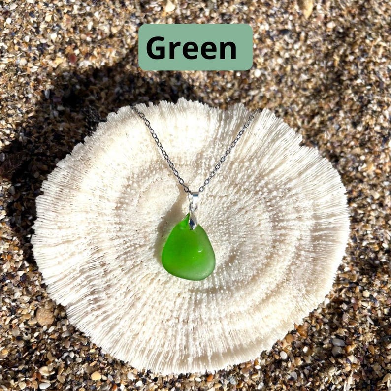 May include: A green sea glass pendant necklace with a silver chain. The pendant is a teardrop shape and is a bright, vibrant green color. The necklace is displayed on a white coral-like surface. The word "Green" is in a green rectangle in the top center.