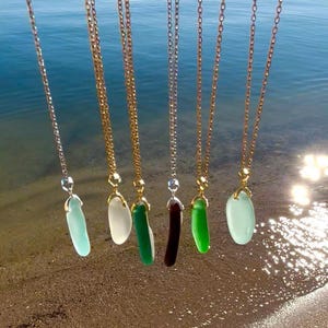 May include: A collection of necklaces with pendants in various colors, including light blue, white, green, and dark red. The pendants are suspended from gold and silver chains, with the ocean in the background.