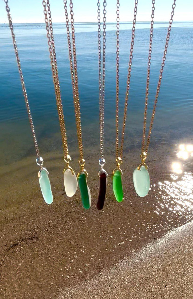 May include: A collection of necklaces with pendants in various colors, including light blue, white, green, and dark red. The pendants are suspended from gold and silver chains, with the ocean in the background.