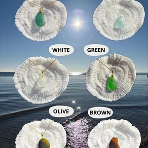 May include: Necklaces with sea glass pendants in various colors, including teal, light aqua, white, green, olive, and brown. Each pendant hangs from a delicate gold or silver chain and rests on a white coral-like base. The background shows a sunny sky and ocean.