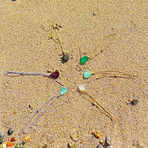 May include: A collection of necklaces with sea glass pendants arranged on a sandy surface. The pendants are various colors, including green, blue, and clear, and are suspended from delicate chains. The necklaces are displayed in a circular pattern.