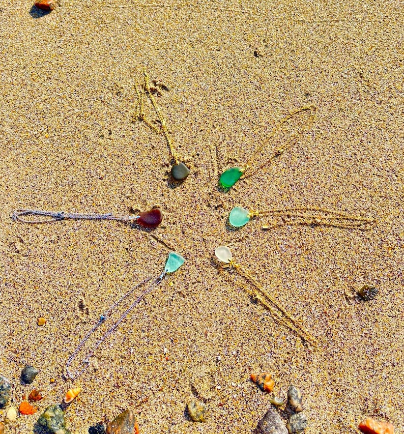 May include: A collection of necklaces with sea glass pendants arranged on a sandy surface. The pendants are various colors, including green, blue, and clear, and are suspended from delicate chains. The necklaces are displayed in a circular pattern.