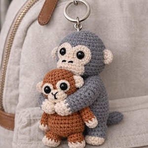 Pode incluir: Um chaveiro de crochê artesanal com duas figuras de macaco. O macaco maior é cinza, abraçando um macaco marrom menor. Ambos têm olhos pretos e rostos e pés creme. O chaveiro está preso a um anel prateado.