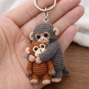 Może przedstawiać: Ręcznie robiony brelok z szydełka przedstawiający dwie obejmujące się małpy. Większa małpa jest szara, a mniejsza brązowa. Brelok ma srebrny metalowy pierścień. Małpy są wykonane z przędzy i mają czarne oczy.